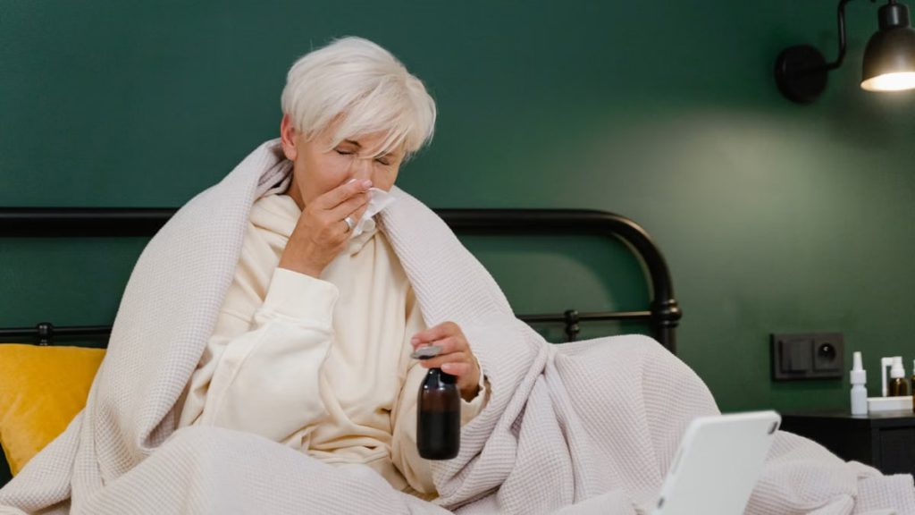 Elderly woman sits in bed feeling unwell, covered with a blanket, holding medicine.